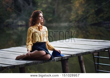 Dreaming girl with curly hair sitting on wooden bridge forest and river on background. Beautiful woman sitting by the lake. Sad girl sitting on bridge. Lonely woman sitting on a wooden bridge. Portrait of young woman on autumn background