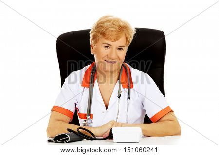 Smile elderly female doctor or nurse sitting behind the desk with bloog preasure gauge