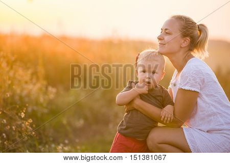 Young beautiful mother hugging her little toddler son on a sunset in a wild flowers field. Happy woman with her baby boy on a summer sunny day. Family walking on the meadow.