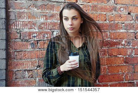 Young sad girl with long hair standing near the brick wall drinking coffee from disposable cup