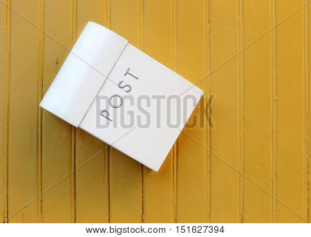 The white mailbox hanging awry on a yellow painted wooden wall.