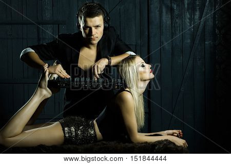 Young man dj in black suit with headset playing music on mixer equipment standing on shoulders and legs of sexy girl blonde in glamour shorts on dark wooden background studio