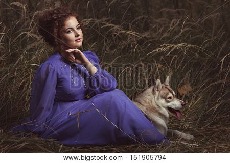 Woman sitting in the tall grass one in a beautiful fairy dress lying beside the dog.