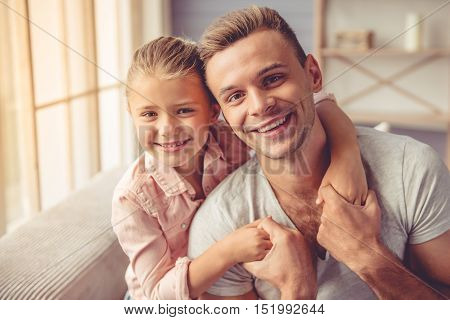 Portrait of handsome young father and his little daughter hugging looking at camera and smiling while sitting on sofa at home