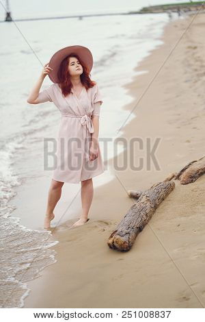 A Young Redheaded Girl In A Large Round Hat And Pink Dress Wallking On The Beach Near Ocean