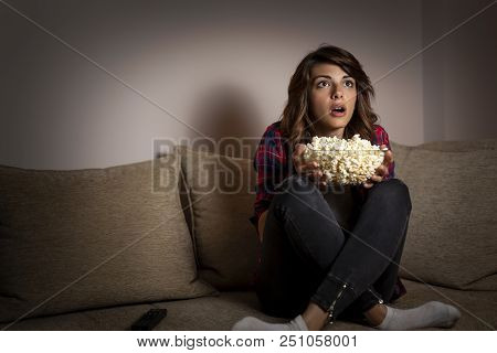 Beautiful Young Woman Sitting In The Dark On A Living Room Couch, Having Fun Watching Tv And Eating 