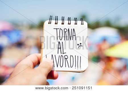 closeup of the hand of a young caucasian man in the seashore of a beach showing a spiral notepad with the text torna al lavoro, back to work in italian, handwritten in it