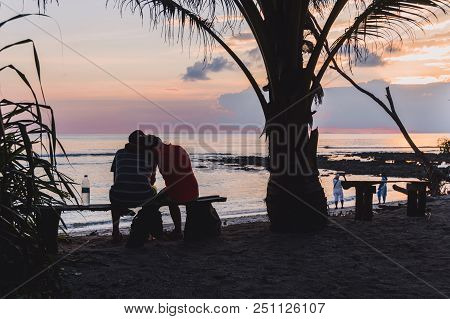 A Young Couple In Love Sits On A Bench And Looks At The Sun Setting Over The Horizon. Silhouettes Of