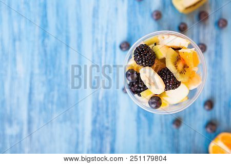 Top View Of A Cup Full Of Delicious Fruits Over A Wooden Desk. Berry, Banana, Kiwi And Grapes Over B