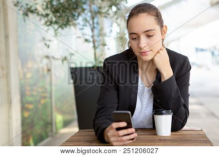 Pensive Female Office Worker Testing New Mobile App During Lunch Break. Beautiful Girl In Jacket Usi
