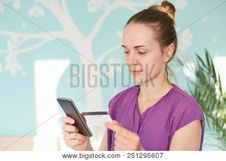 Indoor Shot Of Housewife Holds Cell Phone And Plastic Card, Makes Shopping Online, Chooses What To B