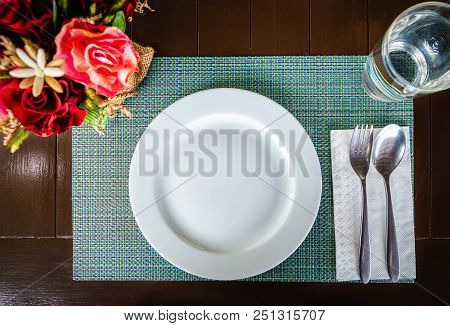 Top View Of White Ceramic Dish With Spoon And Fork On Tablemat On Brown Wooden Table. Glass Of Water