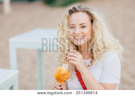 Beautiful Young Smiling Female With Curly Light Hair, Toothy Smile, Recreats On Beach, Drinks Summer