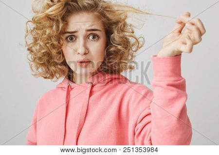 Studio Shot Of Upset Questioned Curly-haired Blonde Woman Pulling Hair Strand That Remained Straight