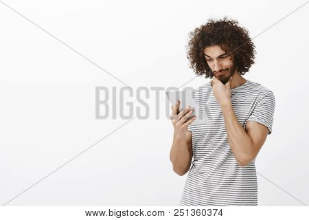 Portrait Of Pleased Curious Attractive Guy With Beard And Afro Haircut, Rubbing Chin While Gazing At
