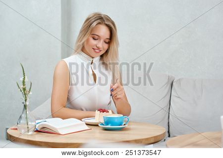 Cute Blonde Girl Eating Raspberry Cake Sitting In Cafe. Keeping , Wearing Beige Blouse, Looking At S