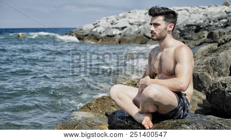 Handsome Shirtless Young Man During Meditation Or Doing An Outdoor Yoga Exercise Sitting On Rocks By
