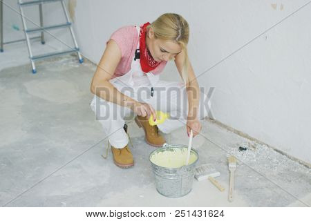 Young blond female in white overalls sitting on hunkers and pouring bright yellow paint creating beautiful pastel colored wall paint in metal bucket with brushes scattered around on floor.