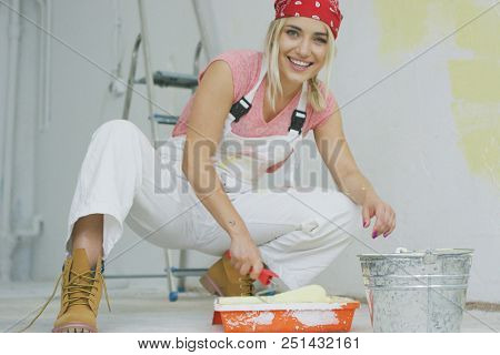 Crop view of happy young attractive woman in white overalls and red bandana sitting on hunkers at bucket and dipping roller in tray with pastel yellow wall paint looking at camera and smiling.