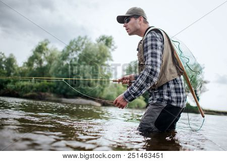 Fisherman In Action. He Stands In Water And Holds Fly Rod In One Hand And Spoon In The Other One. Al
