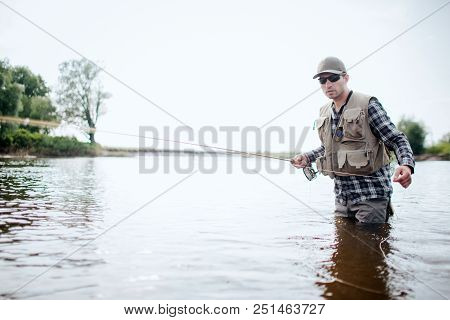 Fisherman In Protection Clothes Is Stnading In Water And Fishing. He Holds Fish Rod In One Hand And 