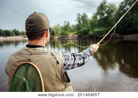 Guy Is Standing And Looking Straight On Water And Trees. He Wears Vest, Cap And Shirt. Also Guy Has 