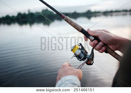 A Picture Of Rotative Spinner That Guy Holds In Hands. He Rotates Reel With Left Hand. He Is At Lake