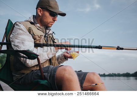 A Picture Of Man Sitting At The Edge Of Water In Folding Chair And Holding Fly Rod. He Has Small Pla