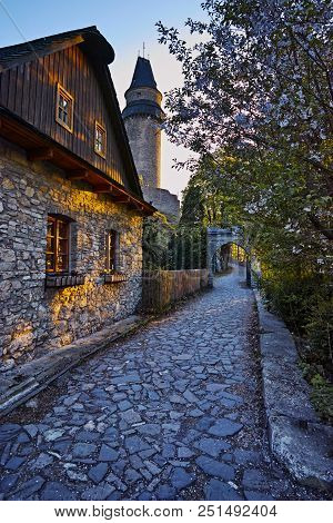 Narrow Street With Timbered House, Old Stone Tower And A White Flowering Tree Illuminated By The Eve