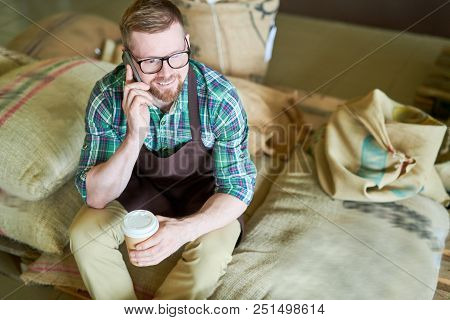 High Angle Portrait Of Handsome Young Man Sitting On Heap Of  Burlap Bags Holding Paper Cup Of Coffe