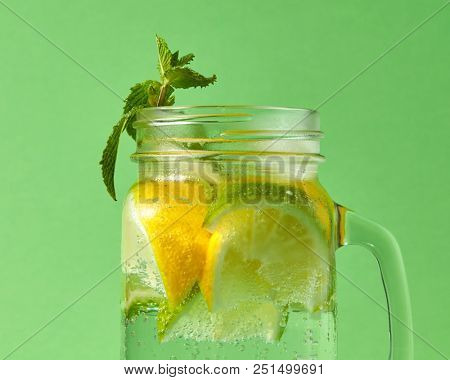 A close-up of green mint and natural citrus fruit-slices of lemon and lime, cubes of ice in a glass jar with a natural cocktail, sparkling bubbles on a green background.