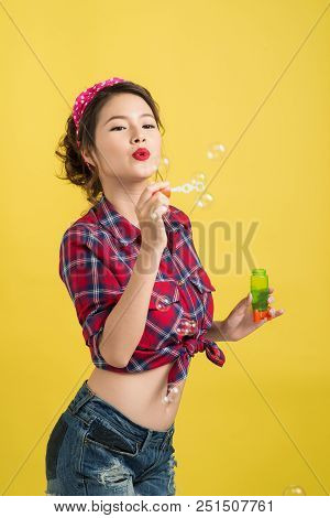 Pin-up Asian Model Blowing Soap Bubbles Over Yellow Background.