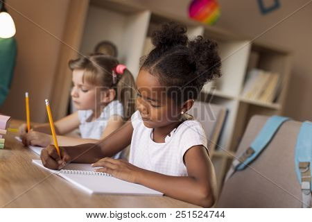 Two Little Girls Sitting At A Desk, Writing In Their Notebooks, Doing A Math Homework And Studying F