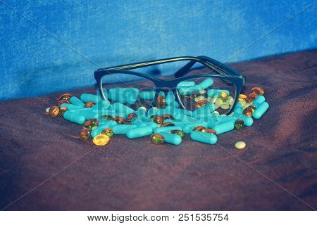 Blue Capsules And Different Pills Are Scattered On The Brown Surface Of The Rowades With Glasses On 