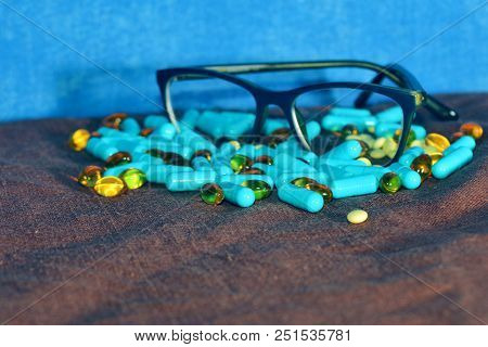 Blue Capsules And Different Pills Are Scattered On The Brown Surface Of The Rowades With Glasses On 