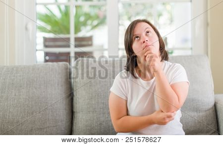 Down syndrome woman sitting on the sofa at home serious face thinking about question, very confused idea