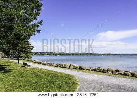 Sidewalk Along The Lake. Rocks Lining The Shoreline Of Canandaigua Lake. View Of The Calm Water And 