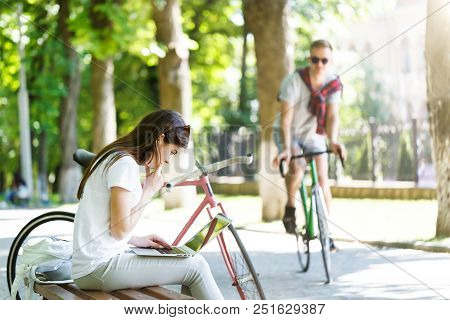Pretty Girl With Bicycle Sitting On Beanch And Using Laptop In The Sunny Summer Park
