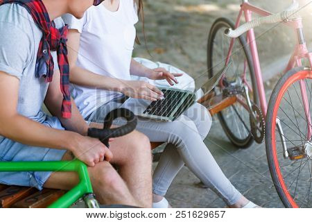 Lovely Couple With Bicycle Sitting On Beanch And Using Laptop In The Sunny Summer Park