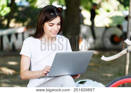 Pretty Girl With Pink Bicycle Sitting On Beanch And Working With Laptop In The Sunny Summer Park
