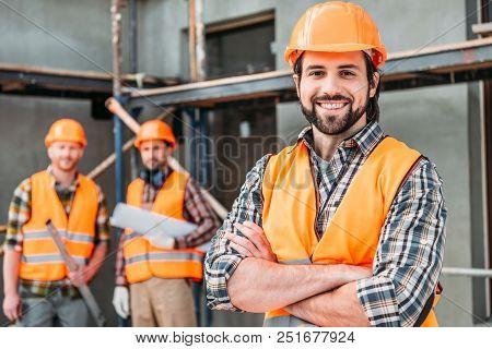 Handsome Smiling Builder Standing At Construction Site With Crossed Arms While His Colleagues Standi