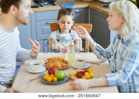 Upset little girl sitting by served table while her parents having quarrel at breakfast
