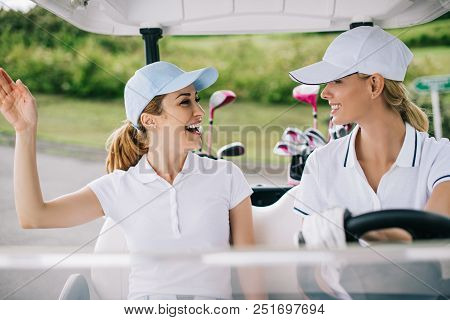 Portrait Of Cheerful Female Golfers In Caps Riding Golf Cart At Golf Course