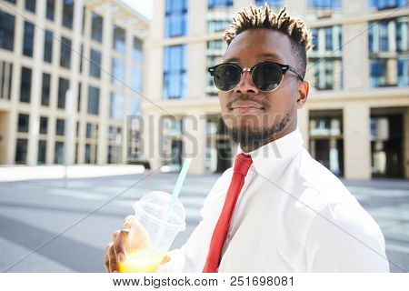 Young African-american man in formalwear and sunglassses having drink in urban environment on summer day
