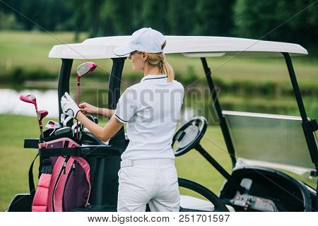 Back View Of Woman In Polo And Cap With Golf Gear Standing At Golf Cart At Golf Course On Summer Day