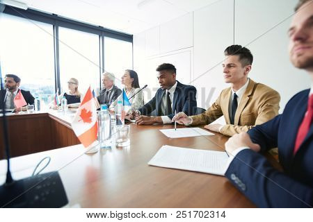 Confident interracial representatives of Commonwealth countries sitting at conference table and listening to rapporteur at meeting