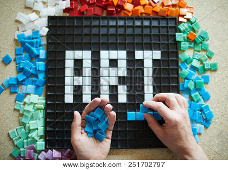 Above view of unrecognizable craftsman laying out  word ART in colored square tiles over black base on table, copy space