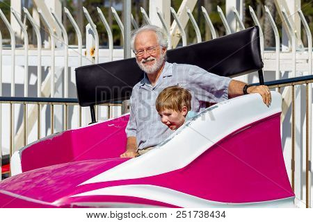 A White Haired Grandfather Looks At The Camera And Smiles As He And His Grandson Ride In A Tilting, 