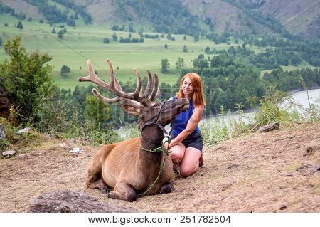 A Young Red-haired Woman Dressed In Sports Clothes Stroking The Horns And Skin Of A Deer Maral