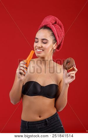 A Smiling Girl Is Eating A Carrot, Prefering It To A Chocolate Donut, Standing On The Red Background